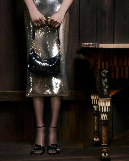 A woman in a sequined dress holds a black Prada handbag, paired with heeled sandals, posing near an ornate wooden table against wood panels.