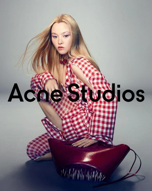 A woman with long blonde hair poses while kneeling in red gingham outfit, with a red sculptural bag, against a grey backdrop.