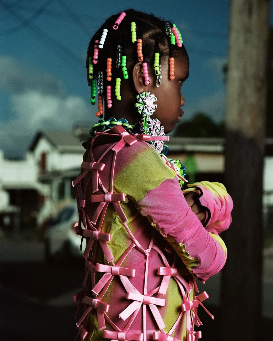 Portrait of a child in colorful dress with bow accents and beaded braids, set against a residential backdrop under a blue sky.