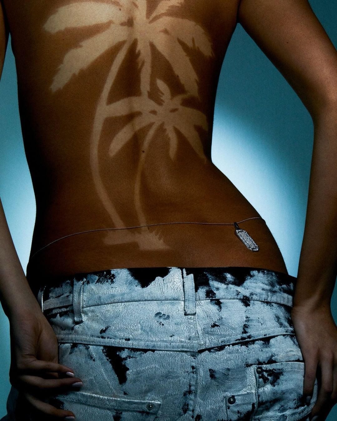 Back view of a woman with tanned palm tree designs on her skin, wearing jeans and a body chain against a blue gradient background.