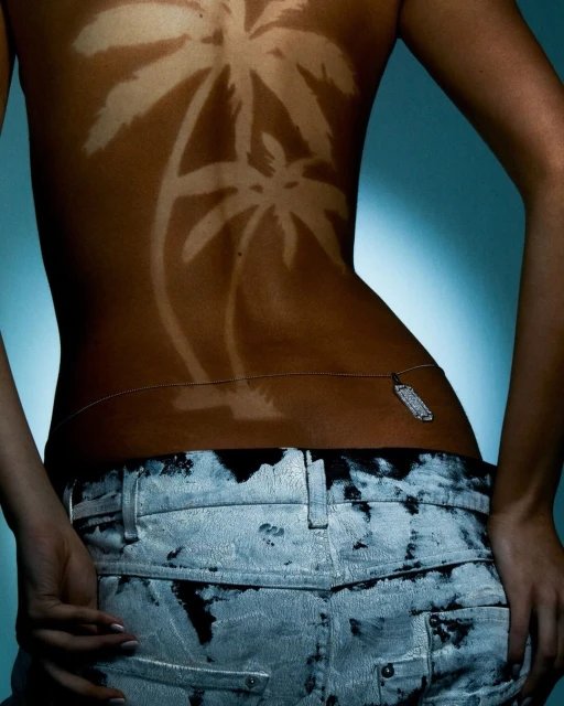 Back view of a woman with tanned palm tree designs on her skin, wearing jeans and a body chain against a blue gradient background.