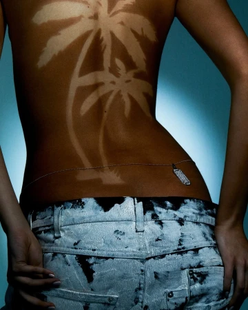 Back view of a woman with tanned palm tree designs on her skin, wearing jeans and a body chain against a blue gradient background.
