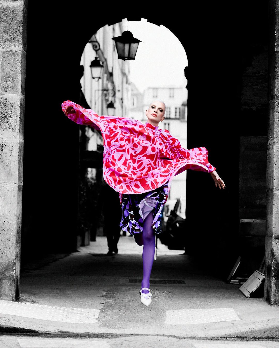 A stylish, bald adult woman is captured mid-stride, wearing a patterned pink top and purple tights in an archway alley.