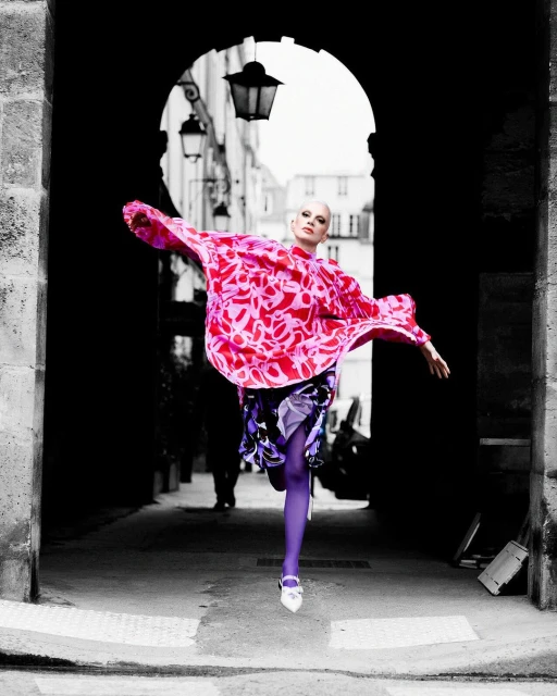 A stylish, bald adult woman is captured mid-stride, wearing a patterned pink top and purple tights in an archway alley.