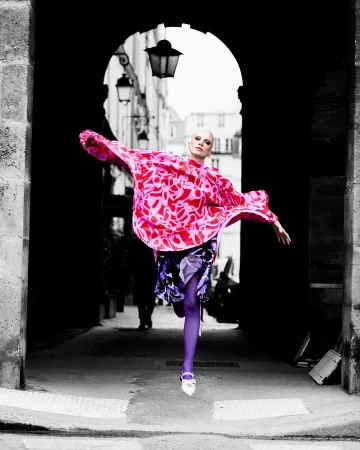 A stylish, bald adult woman is captured mid-stride, wearing a patterned pink top and purple tights in an archway alley.