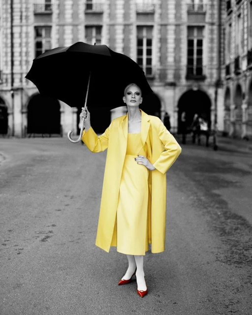 Fashionable woman in yellow dress and coat with black umbrella stands in a gray urban square with a blurred building backdrop.