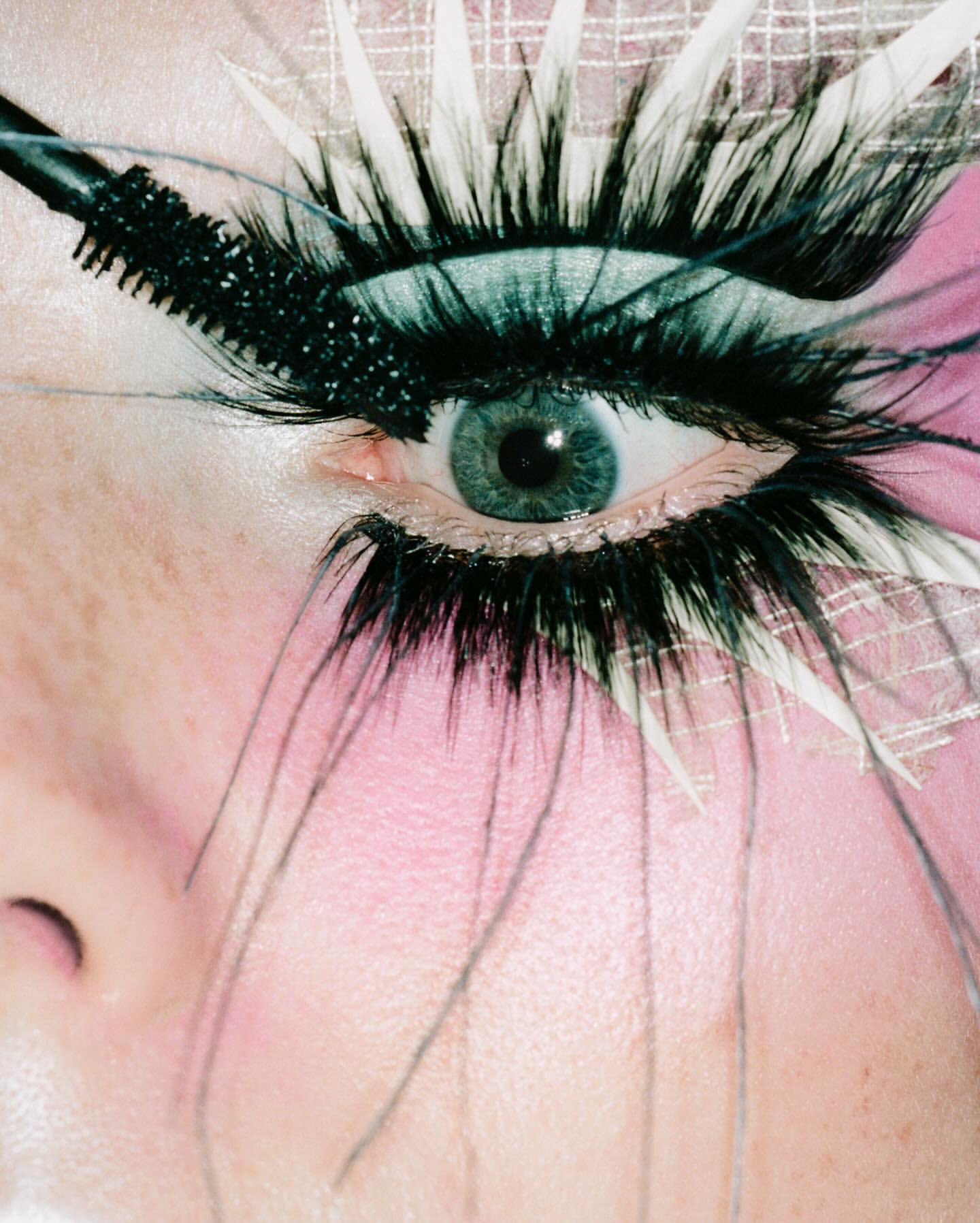 Close-up of a woman's eye with dramatic black eyelashes, mascara wand applying product, fantasy-style makeup, and light pink skin.