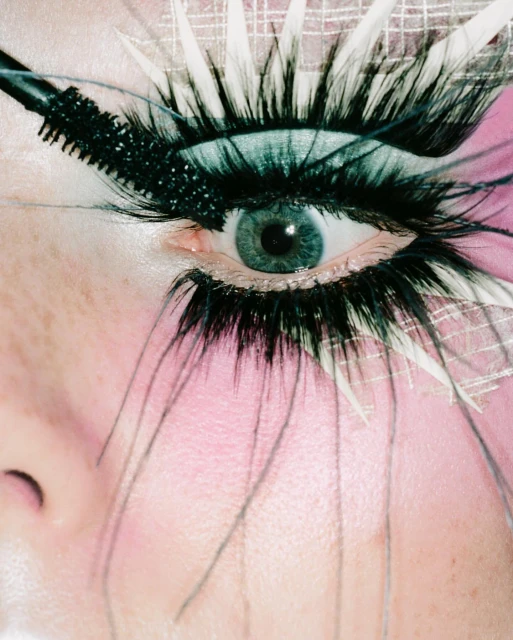 Close-up of a woman's eye with dramatic black eyelashes, mascara wand applying product, fantasy-style makeup, and light pink skin.