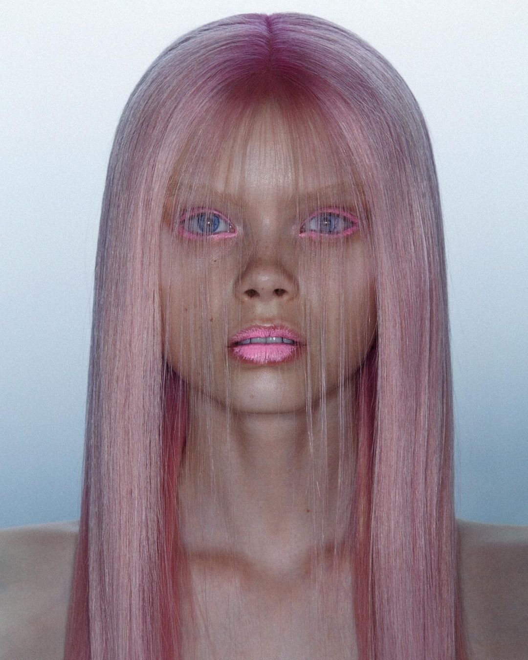 Close-up portrait of an adult woman with pale skin, pink hair, and matching pink makeup against a light gradient background in a photo studio.