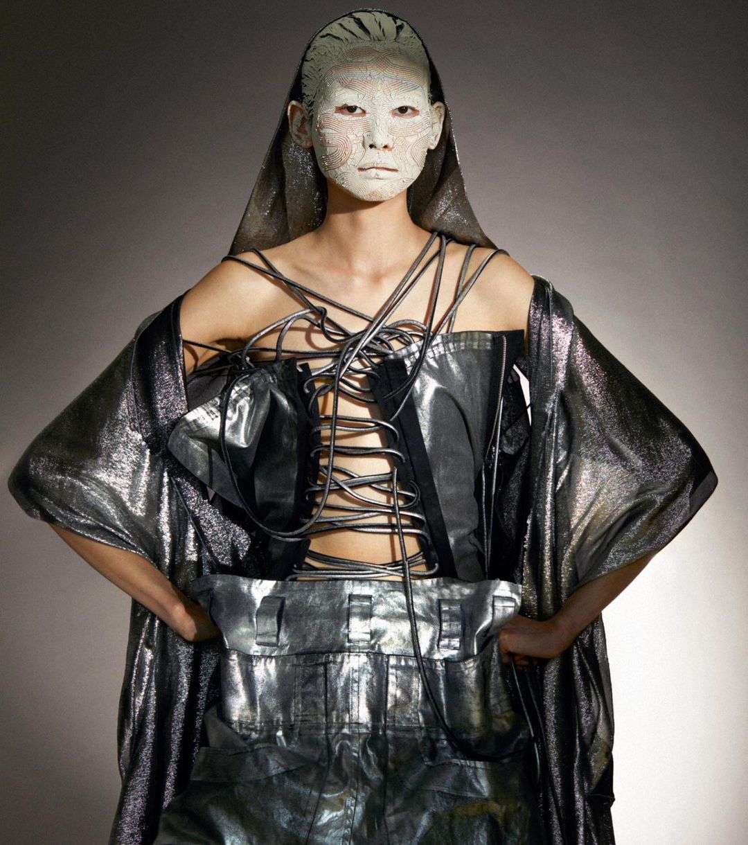 A person in a metallic outfit with geometric face paint and a hood poses with their hands on their waist against a gray background.