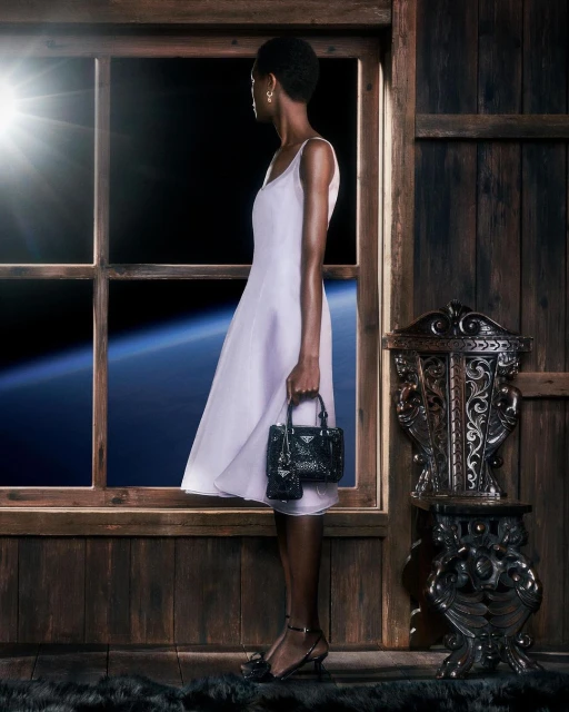 A dark-skinned model in a pale dress holds a black purse by a window with an outer-space view in a wooden-panelled room.