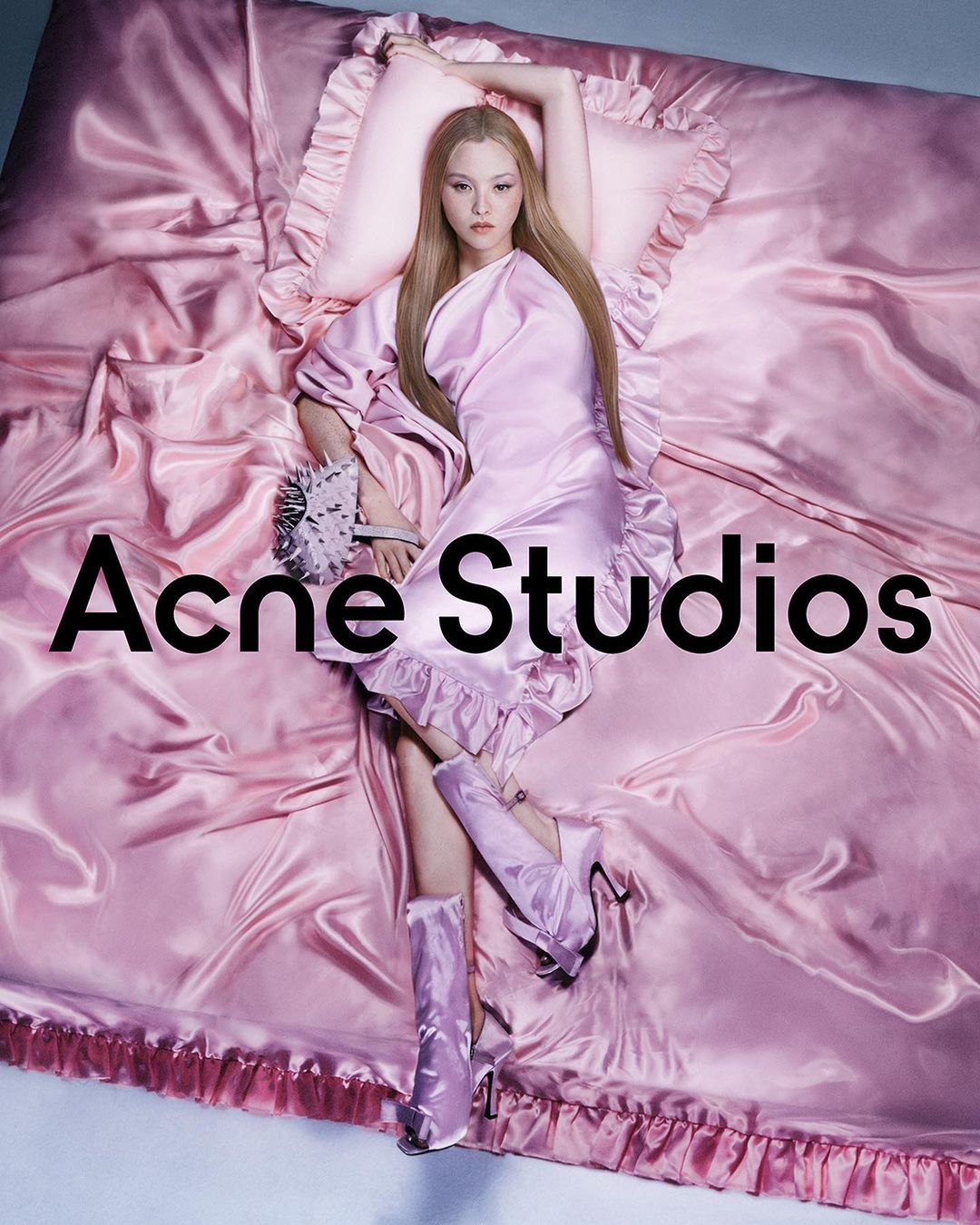 A woman with long blonde hair lies on a pink satin bed with matching pillow and blanket, wearing a pink dress and boots for Acne Studios.