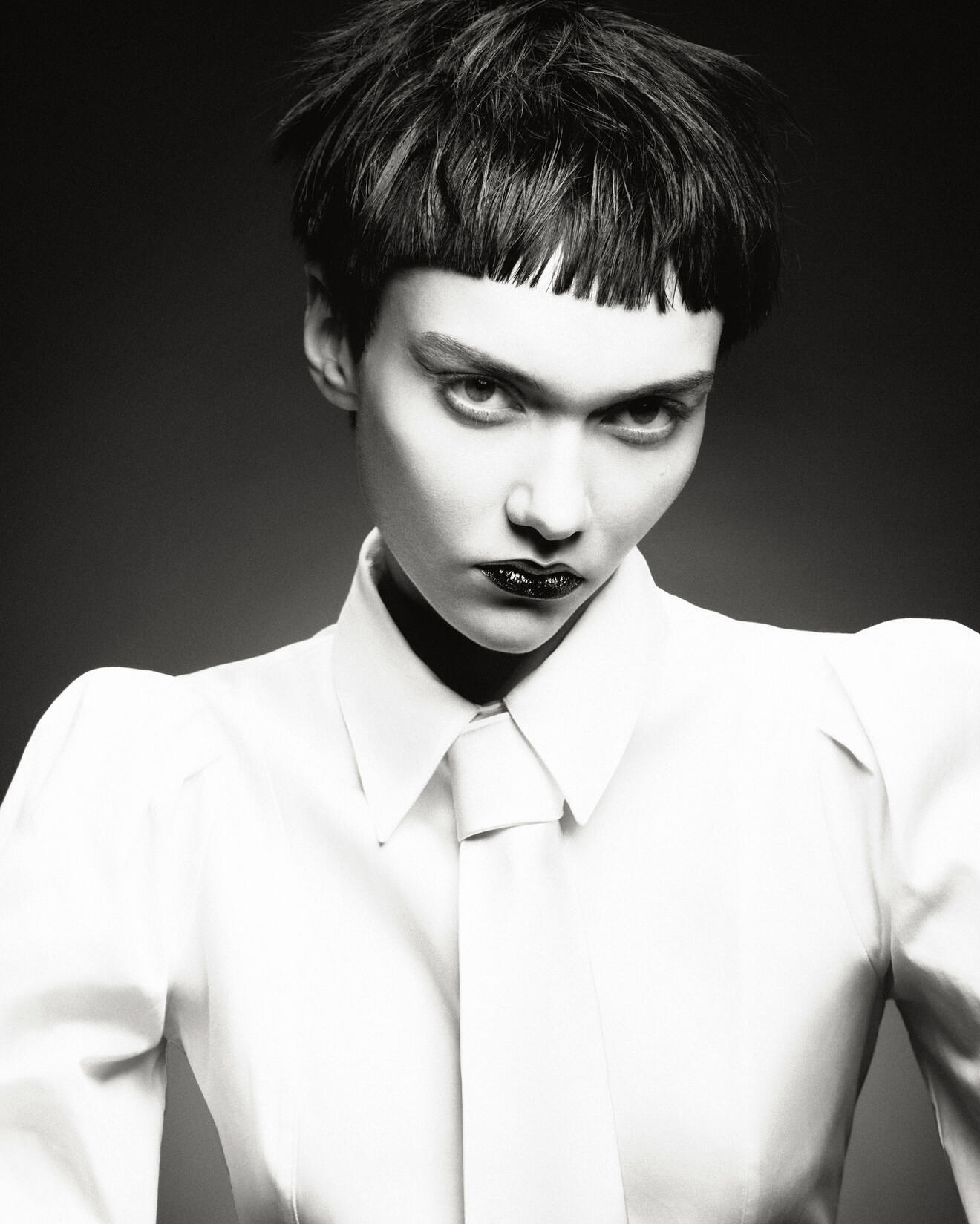 Dramatic black and white studio portrait of an androgynous person with a bowl cut, dark lipstick, and a white shirt and tie.