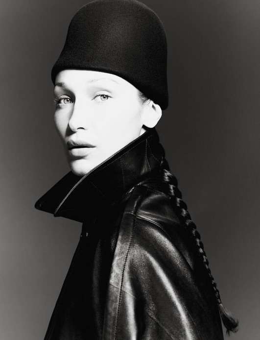 Black and white portrait of a woman with braided hair wearing a black hat and leather jacket against a dark gray backdrop.
