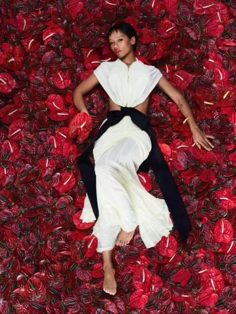 Overhead shot of a woman in a white dress with a black sash lying on a bed of red anthurium flowers in a fashion editorial.