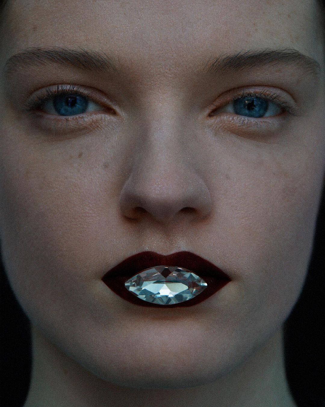 Close-up portrait of a woman with blue eyes and dark lipstick, featuring a jewel-toned gem accenting her lips against a dark background.