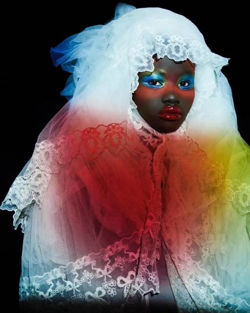Dark-skinned model in lace dress with bold blue and red makeup, posing against a dark background, wearing an elaborate white lace veil.