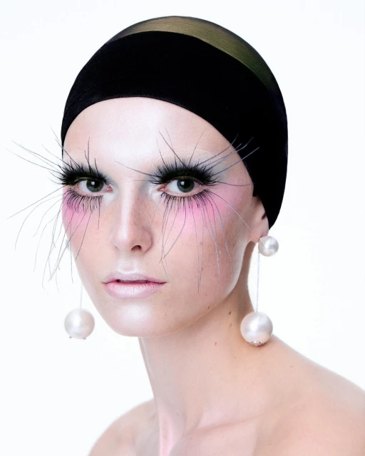 Close-up of a model with a black head covering, pink makeup, and large pearl earrings against a bright white background in a beauty shot.