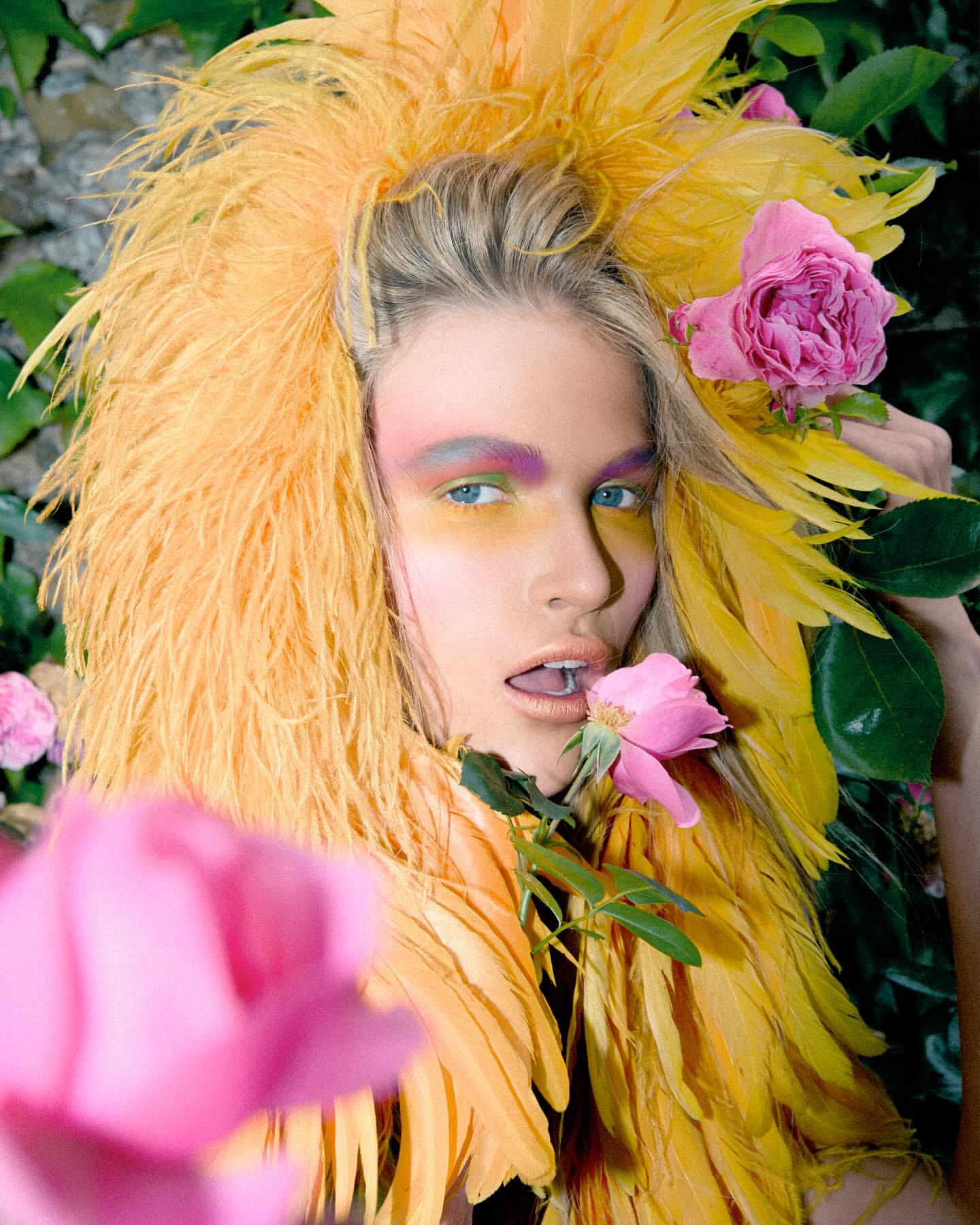Woman with yellow feather headdress, artistic makeup, and pink roses. Close-up fashion beauty shot in an outdoor setting.