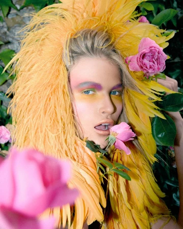 Woman with yellow feather headdress, artistic makeup, and pink roses. Close-up fashion beauty shot in an outdoor setting.