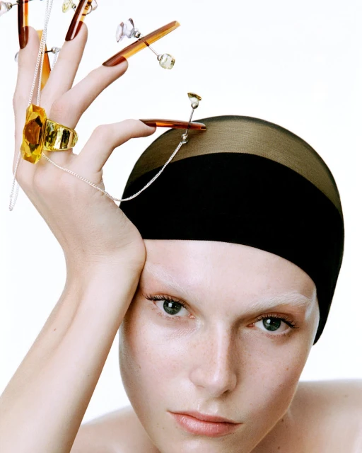 Close-up studio shot of a fair-skinned model with bleached eyebrows, wearing a black cap and gold jewelry, posing against a white backdrop.