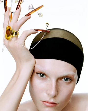 Close-up studio shot of a fair-skinned model with bleached eyebrows, wearing a black cap and gold jewelry, posing against a white backdrop.