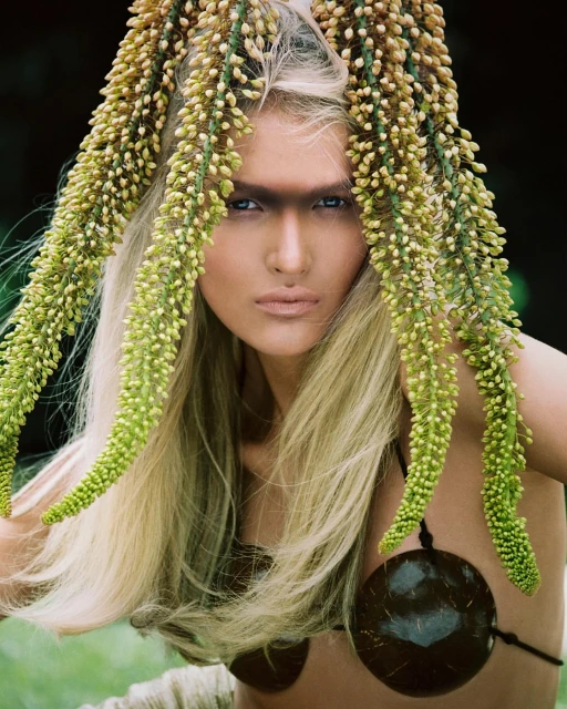 Portrait of a young woman with blond hair and dramatic makeup, adorned with greenery and wearing a coconut shell top against a blurred background.