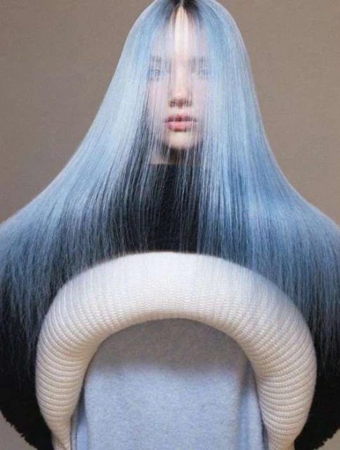 A fashionable woman with dyed blue hair poses indoors, holding a cushioned neck support. The scene is simple with a neutral background.