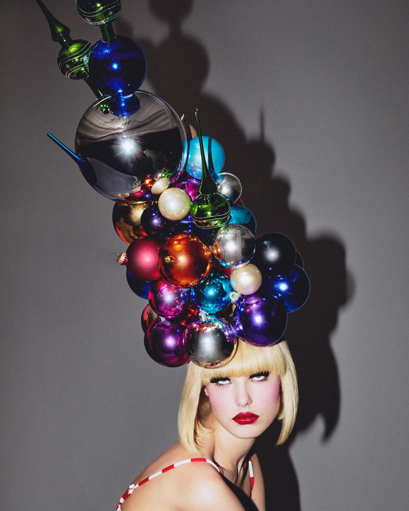 Woman with blonde hair and red lipstick wearing a hat made of colorful Christmas ornaments against a solid gray background. 