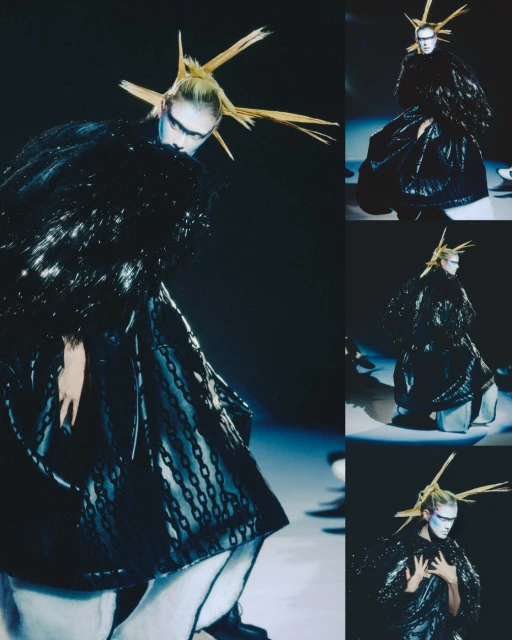 Runway fashion show featuring a model in an elaborate, dark couture gown with avant-garde styling and bold makeup against a dark background.