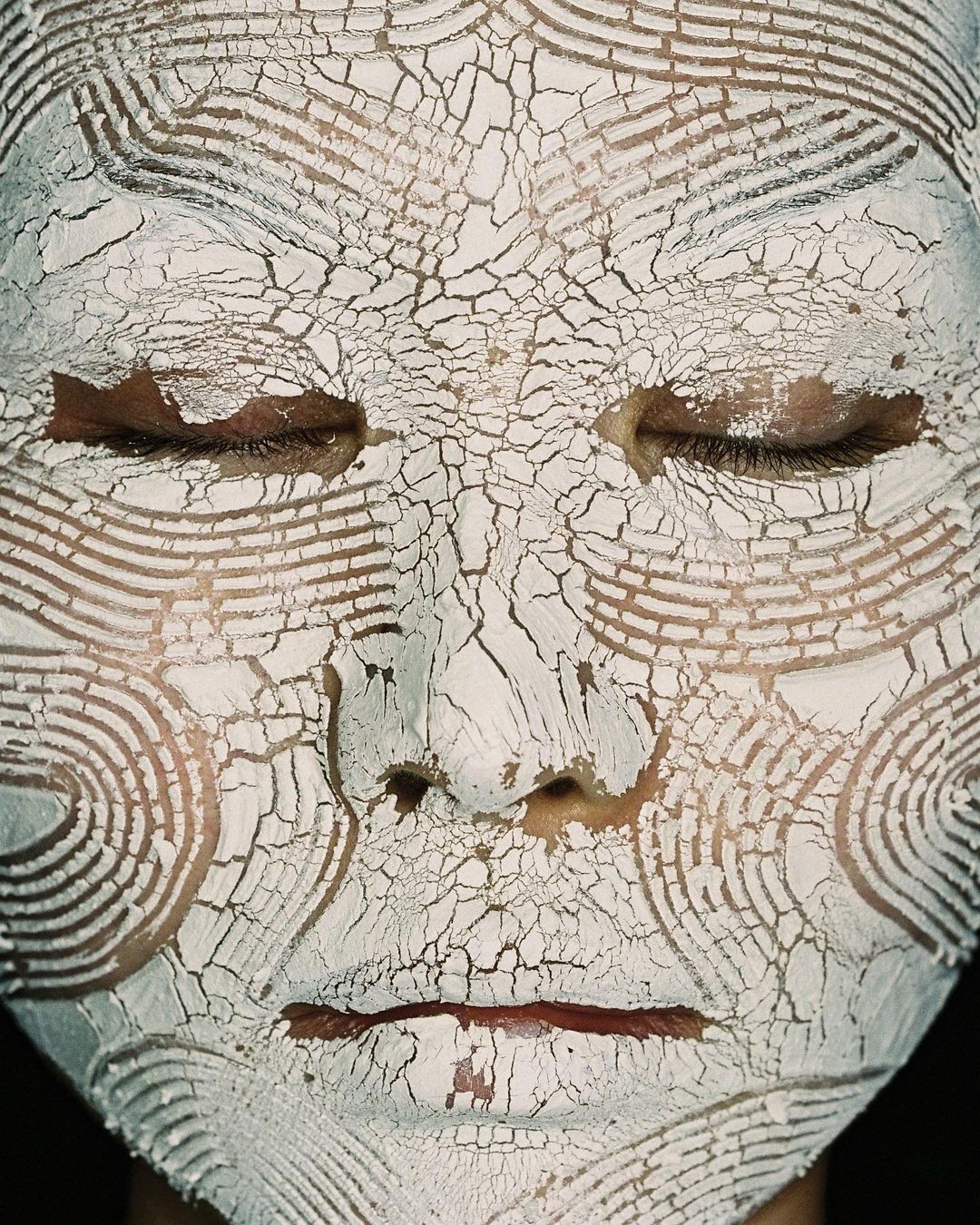Close-up portrait of a person's face covered in a cracked, white clay-like mask with patterned concentric lines around the face.