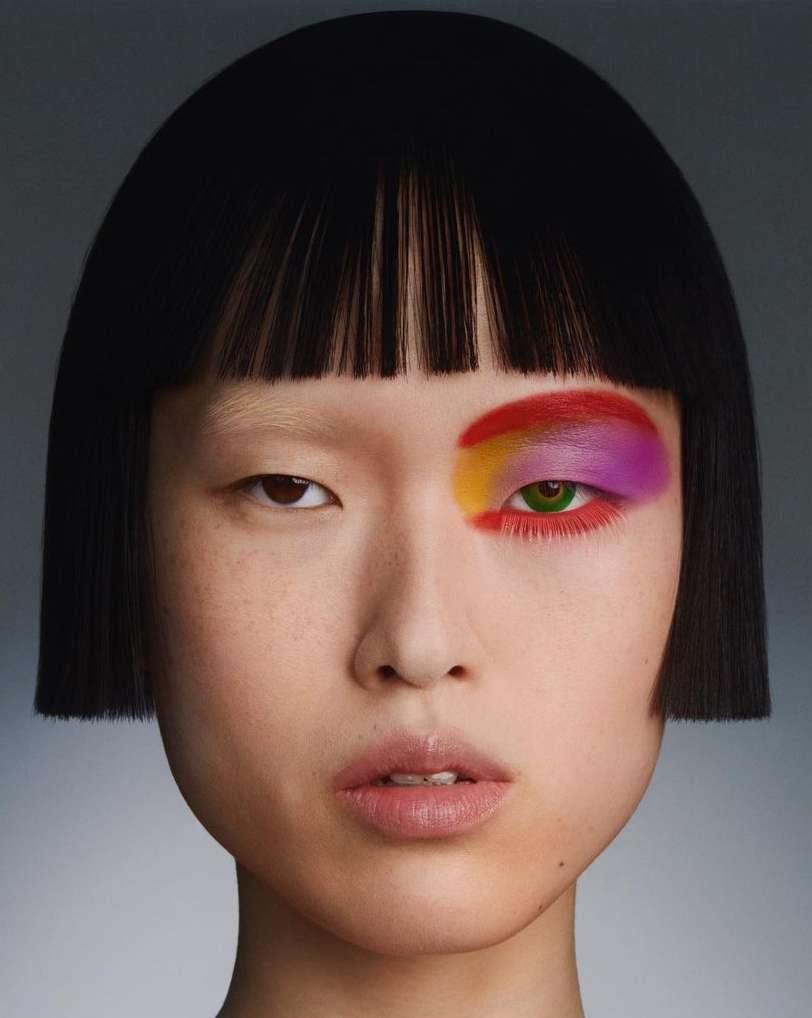 Close-up portrait of a woman with colorful eyeshadow and a short, straight black bob hairstyle against a neutral gray background.