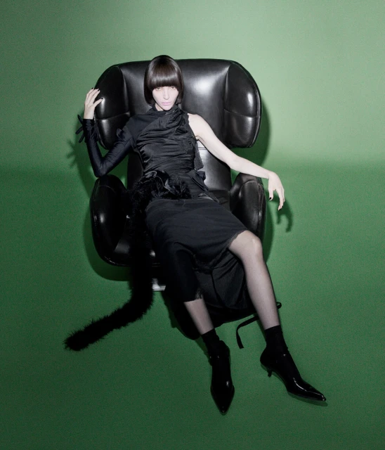 A fashion shot of a woman with a blunt haircut posing in a sculptural black chair against a vibrant green backdrop in a photo studio.