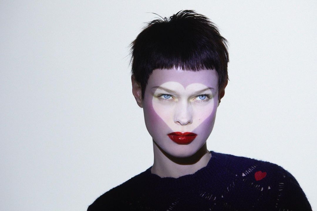 Close-up portrait of an adult woman with short hair wearing dark knitwear, with a heart-shaped light pattern projected on her face.