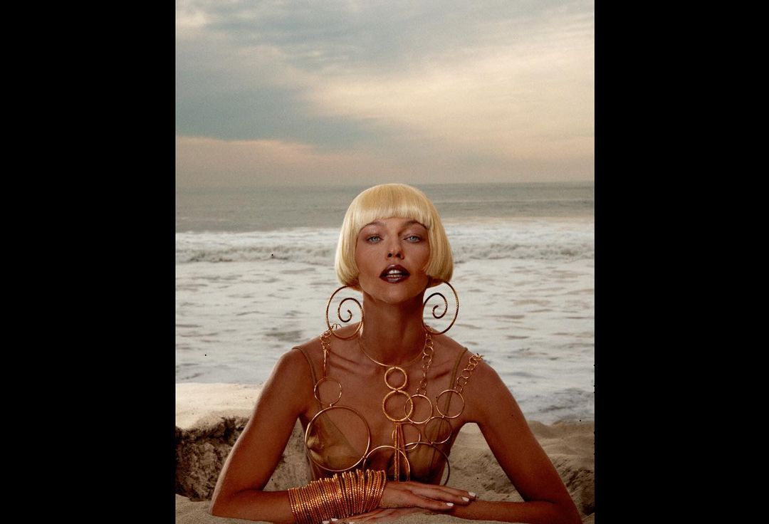 Woman on beach with blonde bob hairstyle, wearing statement jewelry. Ocean backdrop with sandy beach and sky. Beauty fashion shot.