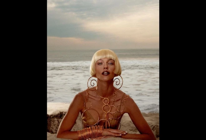 Woman on beach with blonde bob hairstyle, wearing statement jewelry. Ocean backdrop with sandy beach and sky. Beauty fashion shot.