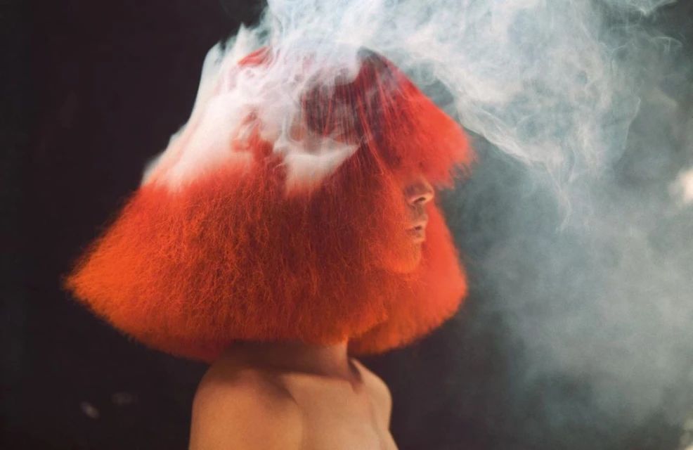 Adult in profile wearing an orange wig with fog against a dark background in a beauty shot, dramatic lighting, and high contrast.