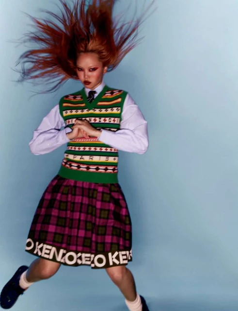 Young adult woman with red hair in motion poses in a school-inspired fashion outfit against a light blue background, in a studio shot.