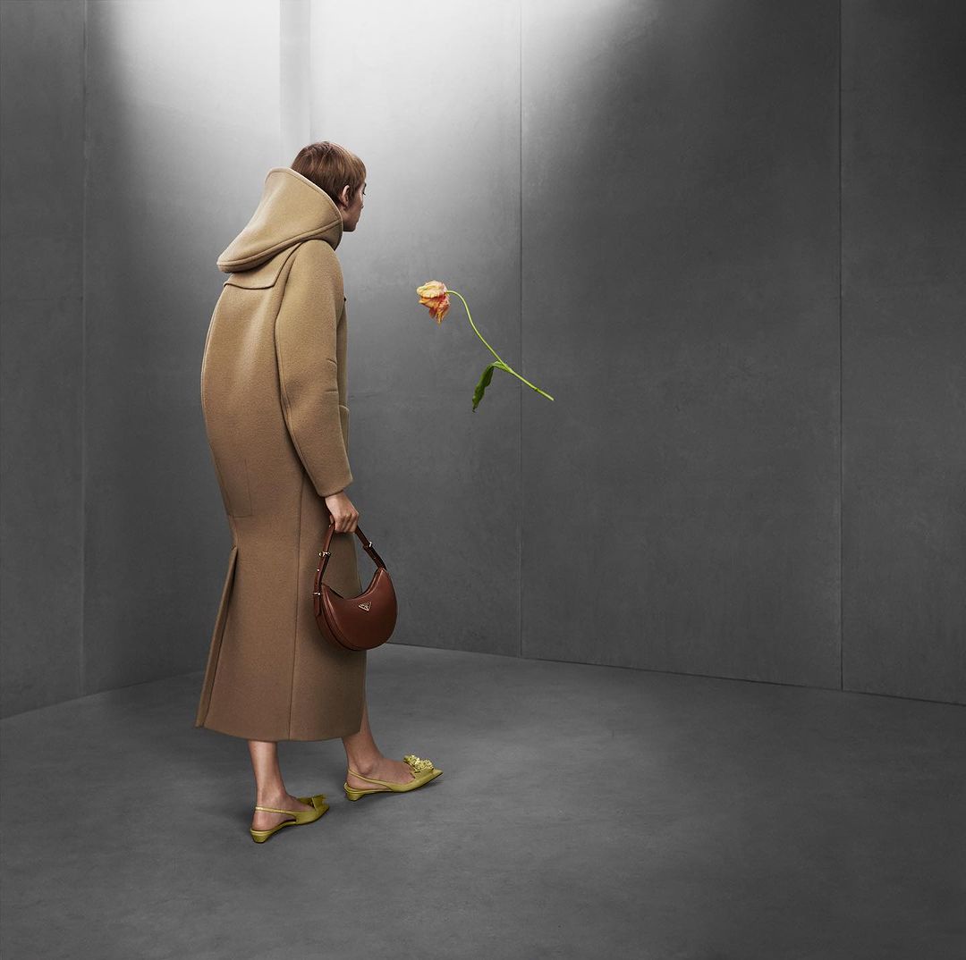 Woman in a tan coat holds a brown purse and looks at a flower in a grey room, a fashion shot.