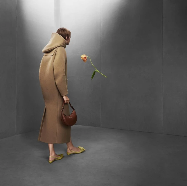 Woman in a tan coat holds a brown purse and looks at a flower in a grey room, a fashion shot.