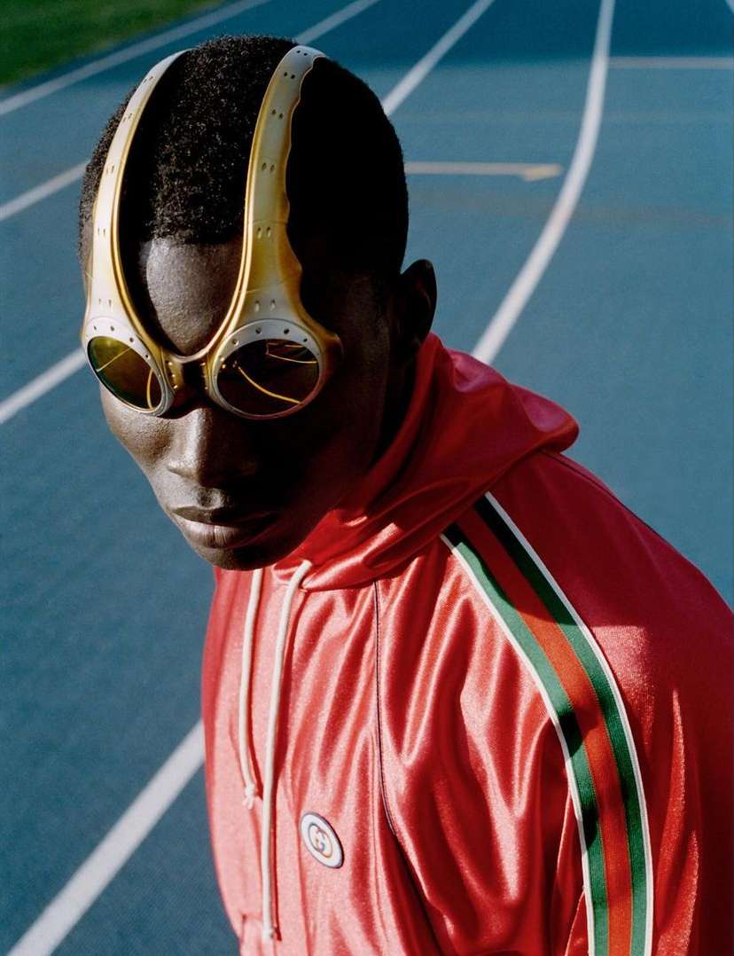 Portrait of a black male model wearing futuristic eyewear and a red tracksuit with green and black stripes on a blue track.