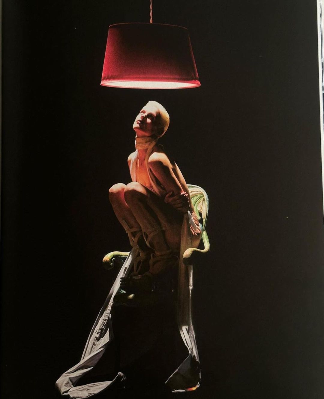 Striking image of a woman with short blonde hair posing on an ornate chair under a red pendant lamp, against a stark black backdrop.