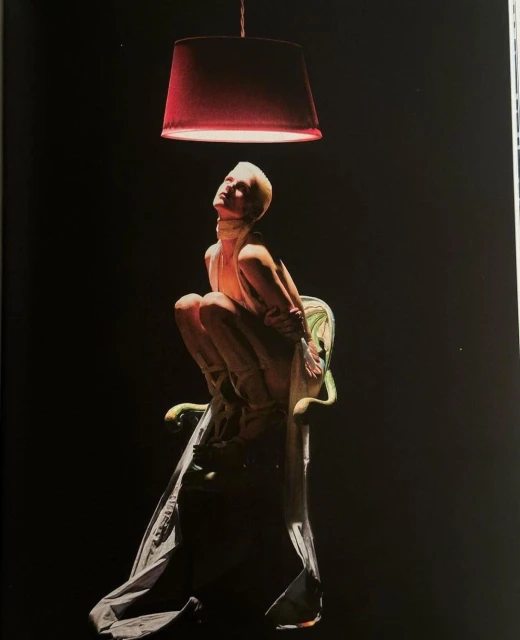 Striking image of a woman with short blonde hair posing on an ornate chair under a red pendant lamp, against a stark black backdrop.