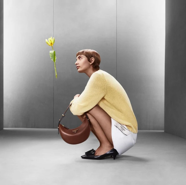 Woman crouches indoors with a Prada purse, glancing up at a yellow flower; she wears a yellow sweater and black shoes, against a grey backdrop.