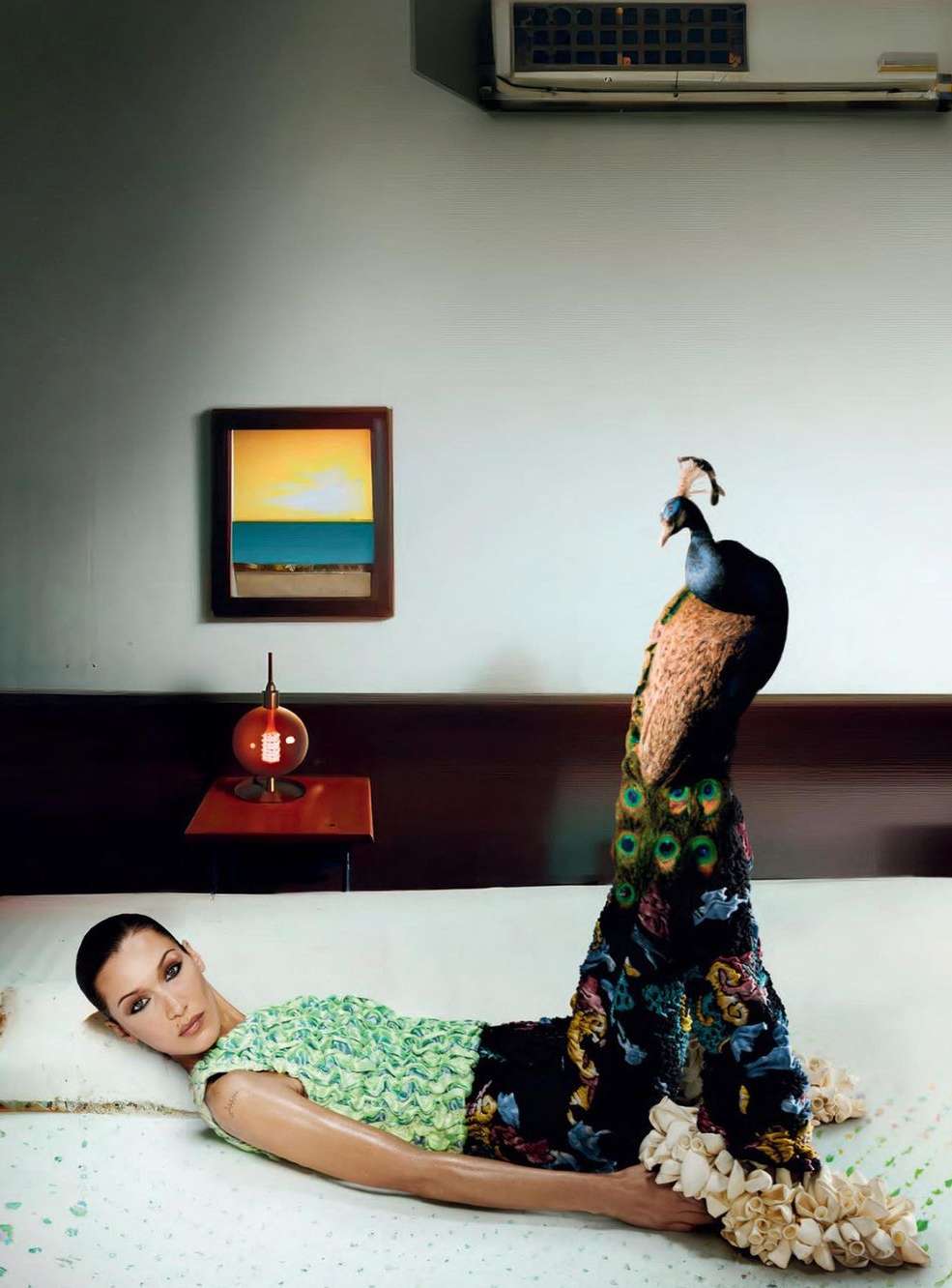 Adult female model laying on a bed with a peacock standing near her legs in a room with a lamp and a framed picture.