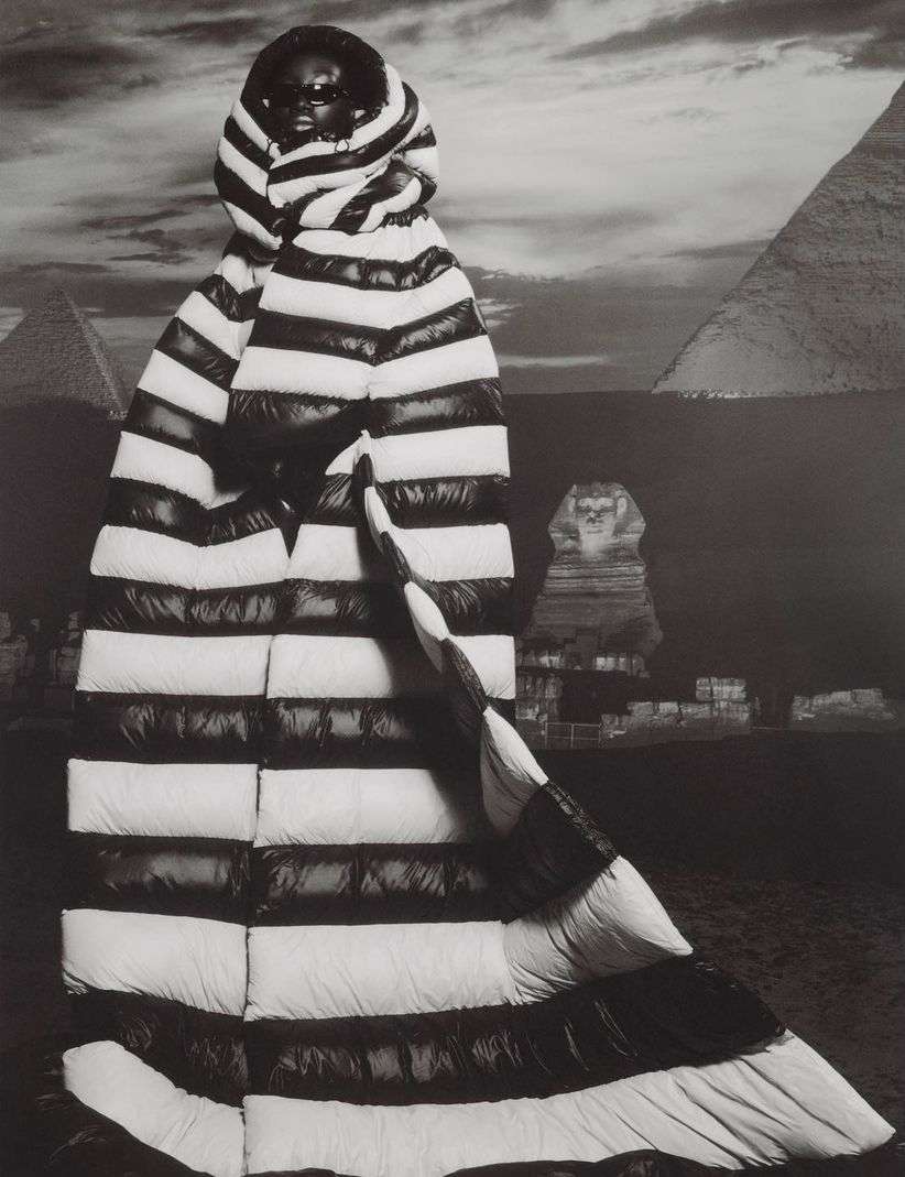 A striking black and white portrait of a person in a striped puffer coat, posing in front of an Egyptian pyramid and sphinx backdrop.