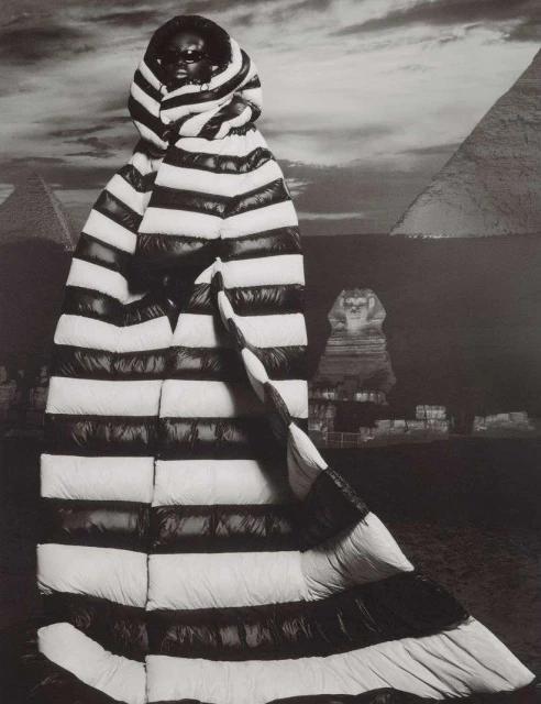 A striking black and white portrait of a person in a striped puffer coat, posing in front of an Egyptian pyramid and sphinx backdrop.
