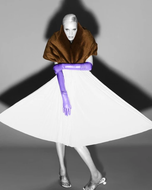 Fashionable adult female model posing in a white dress, brown shawl, and purple gloves, set against a grey background with dramatic lighting.