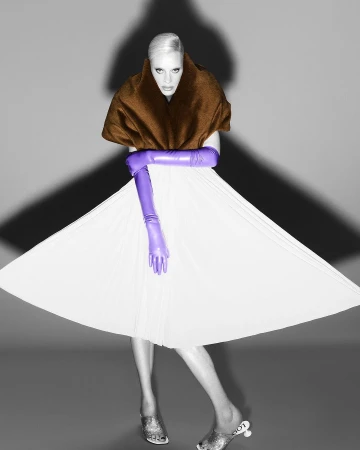 Fashionable adult female model posing in a white dress, brown shawl, and purple gloves, set against a grey background with dramatic lighting.