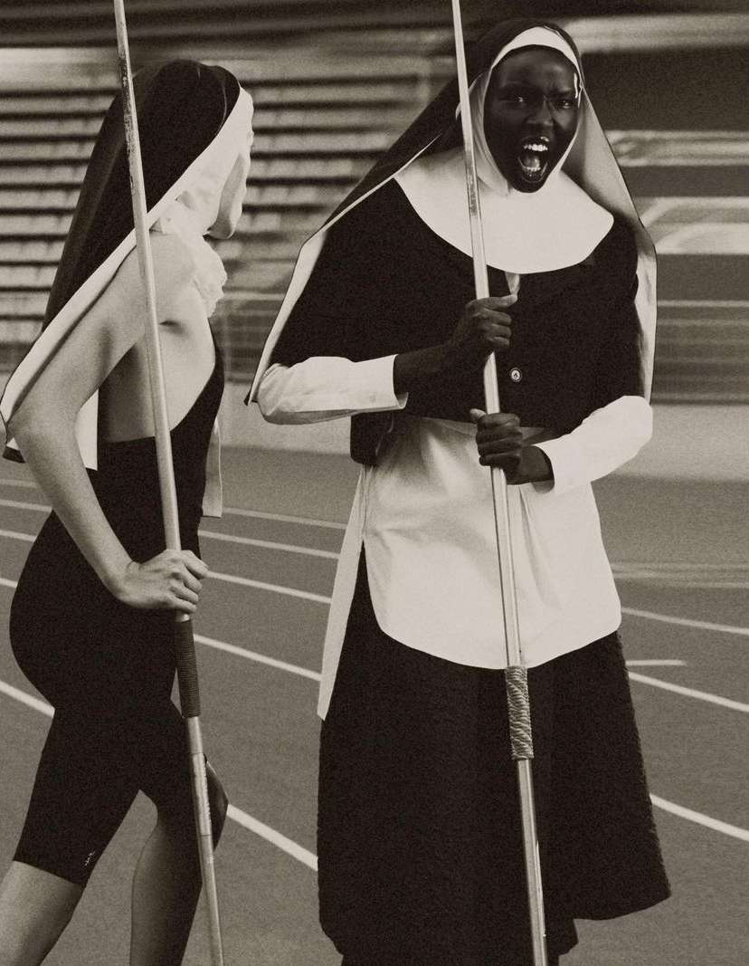 Two adult women in religious nun costumes on a running track, one with an open mouth, holding javelins, indoor setting.